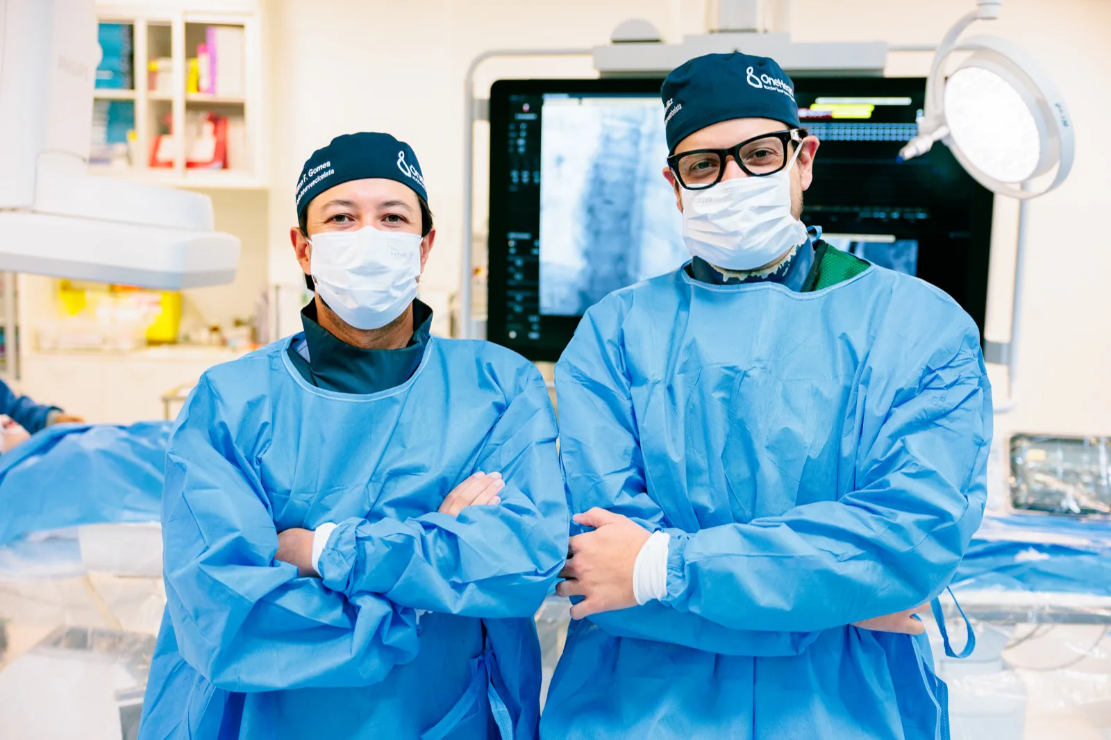 Dois cardiologistas da equipe OneHeart paramentados no Centro de Hemodinâmica, com o monitor de angiografia ao fundo